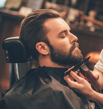 Man in barber chair having beard trimmed by barber with clippers and comb